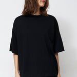 A woman stands in a studio wearing an oversized black short-sleeve top, black pants, and black pointed shoes against a plain white background.