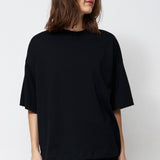 A woman stands in a studio wearing an oversized black short-sleeve top, black pants, and black pointed shoes against a plain white background.