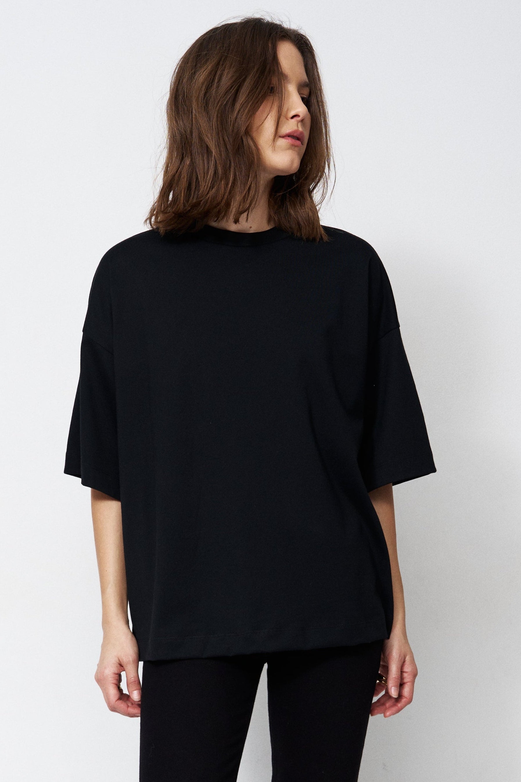 A woman stands in a studio wearing an oversized black short-sleeve top, black pants, and black pointed shoes against a plain white background.