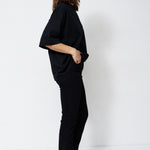A woman stands in profile wearing a black short-sleeve top and black pants against a plain white studio background.