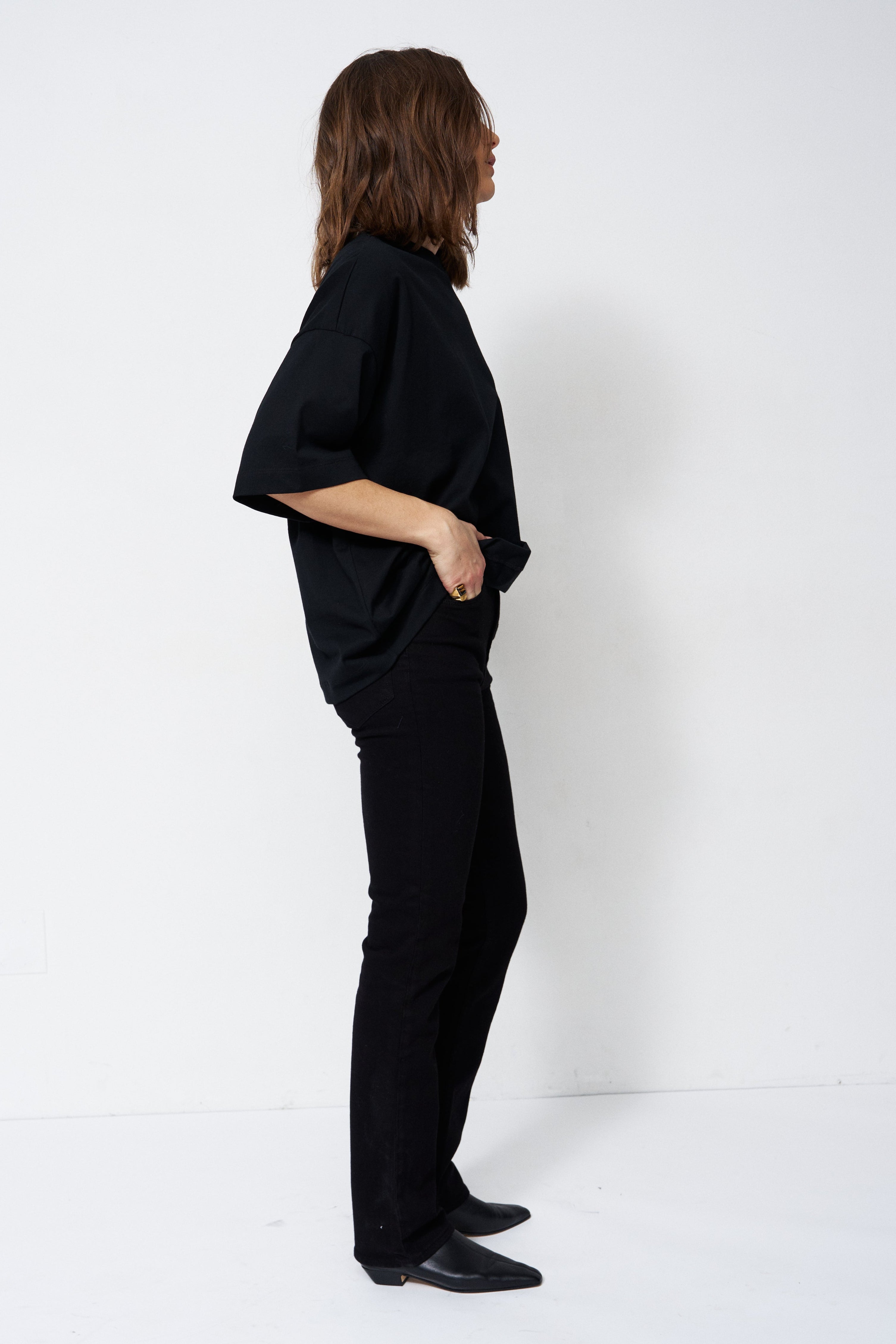 A woman stands in profile wearing a black short-sleeve top and black pants against a plain white studio background.