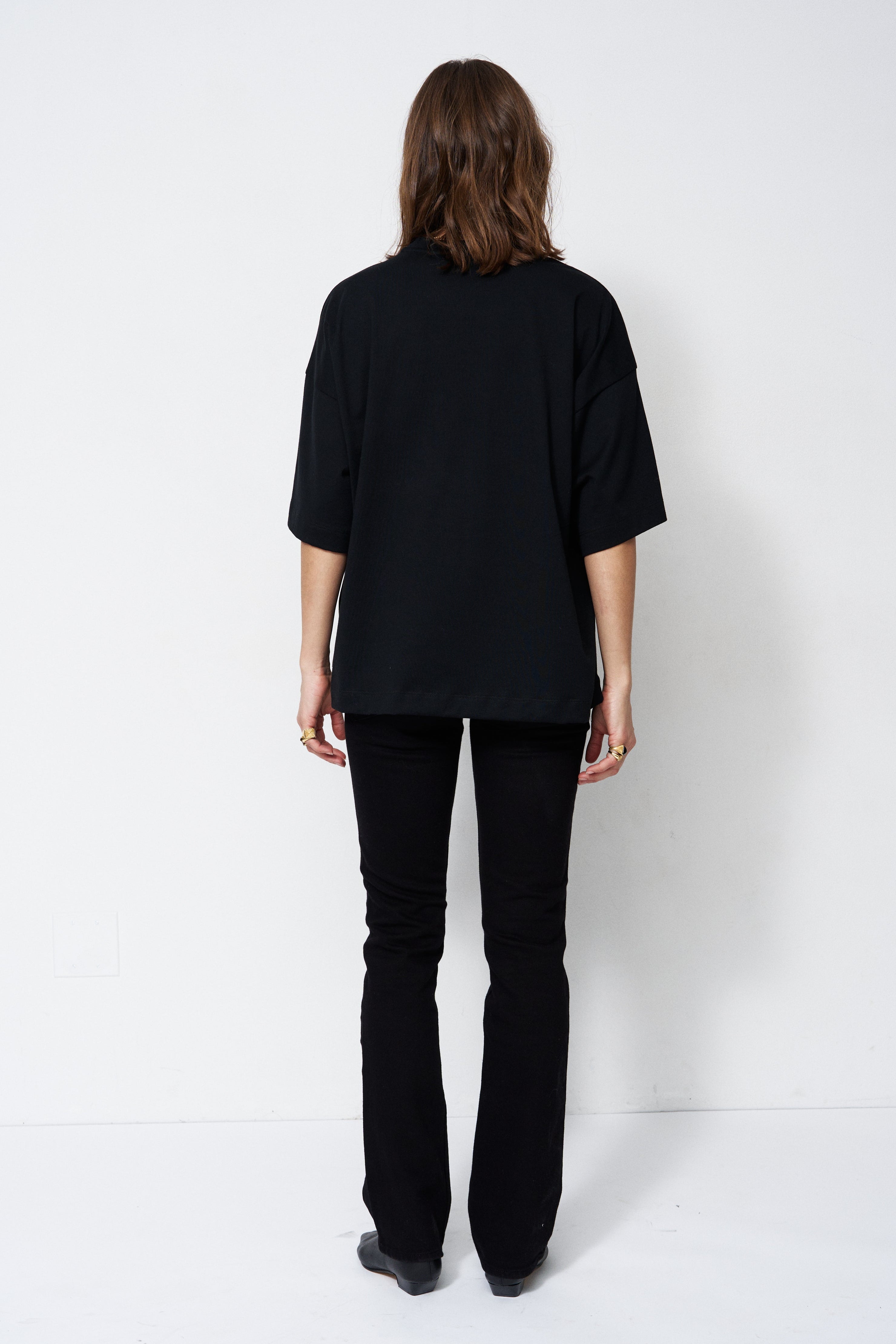A model viewed from the back wearing an oversized black short-sleeve top and black flared pants against a plain white studio background.