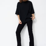 A woman modeling a black oversized short-sleeve mock-neck top with black pants and black shoes, standing against a plain white studio background.