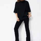 A woman modeling a black oversized short-sleeve mock-neck top with black pants and black shoes, standing against a plain white studio background.