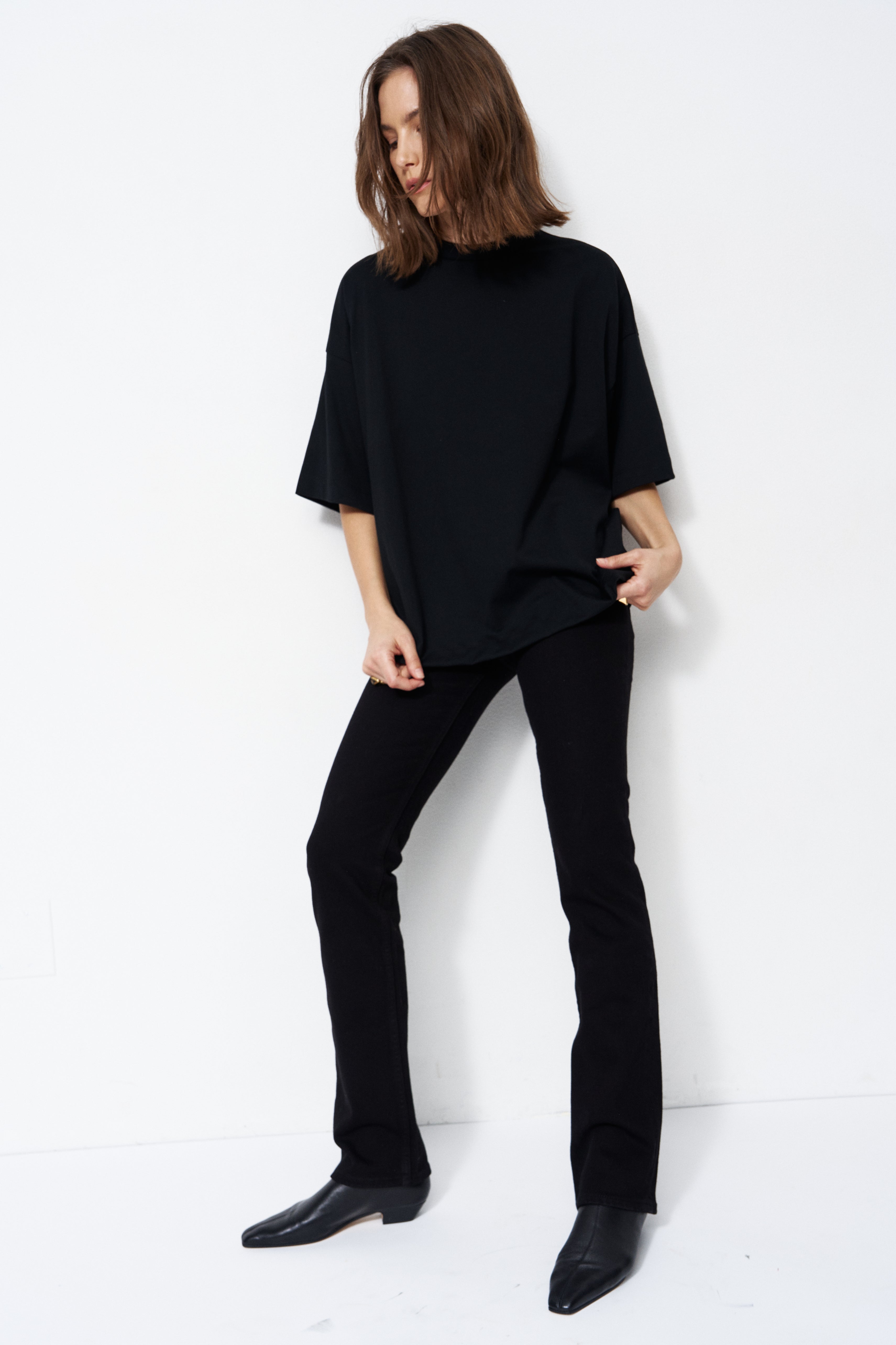 A woman modeling a black oversized short-sleeve mock-neck top with black pants and black shoes, standing against a plain white studio background.