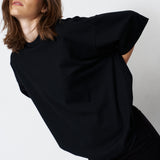 A woman wearing an oversized black short-sleeve top and black pants, posed against a plain light gray studio background.