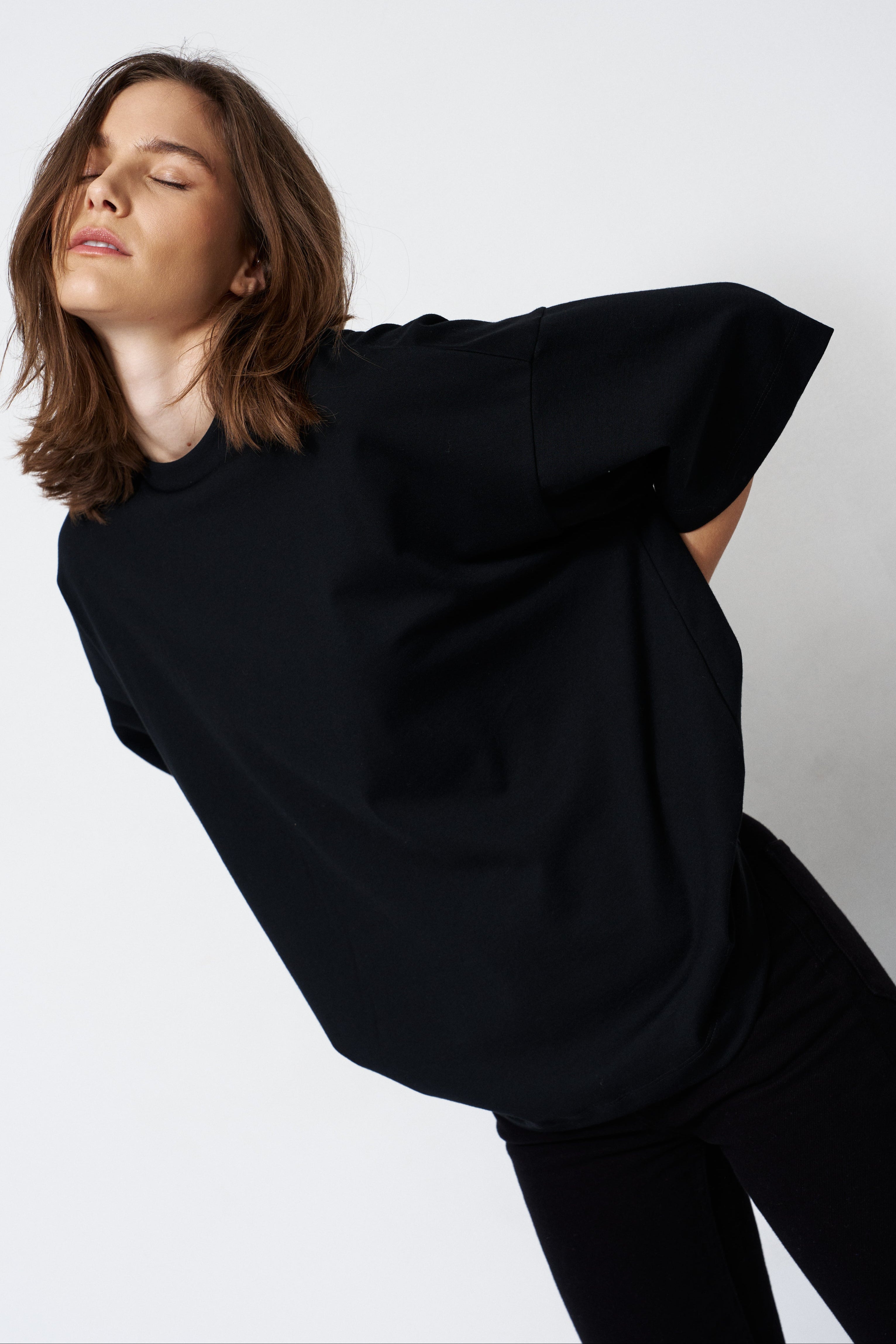 A woman wearing an oversized black short-sleeve top and black pants, posed against a plain light gray studio background.