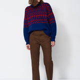 A woman stands against a plain white studio background wearing a navy and red patterned knit sweater, brown trousers, and dark ankle boots.