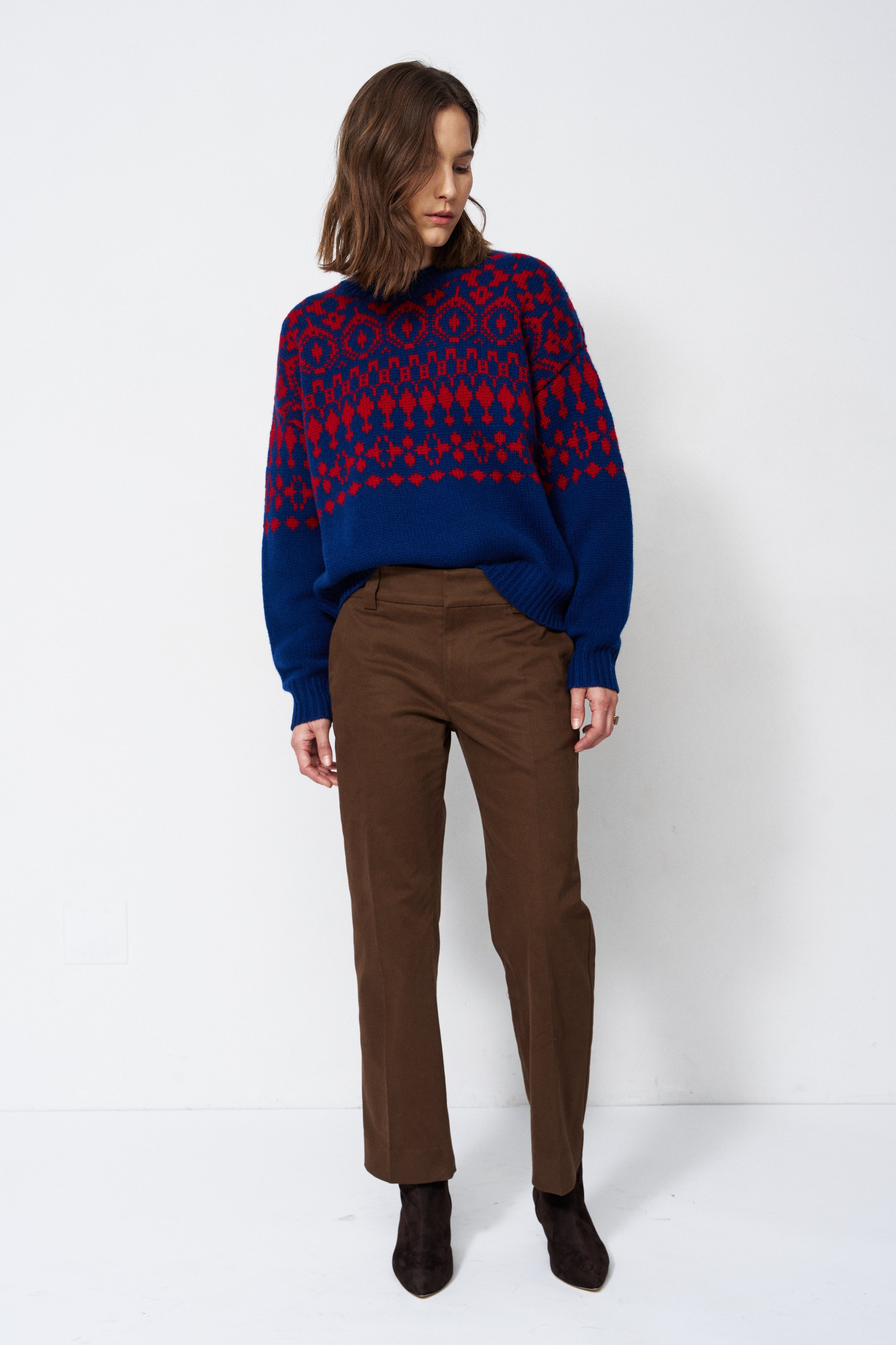 A woman stands against a plain white studio background wearing a navy and red patterned knit sweater, brown trousers, and dark ankle boots.