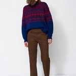 A woman stands against a plain white studio background wearing a navy and red patterned knit sweater, brown trousers, and dark ankle boots.