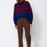 A woman stands against a plain white studio background wearing a navy and red patterned knit sweater, brown trousers, and dark ankle boots.
