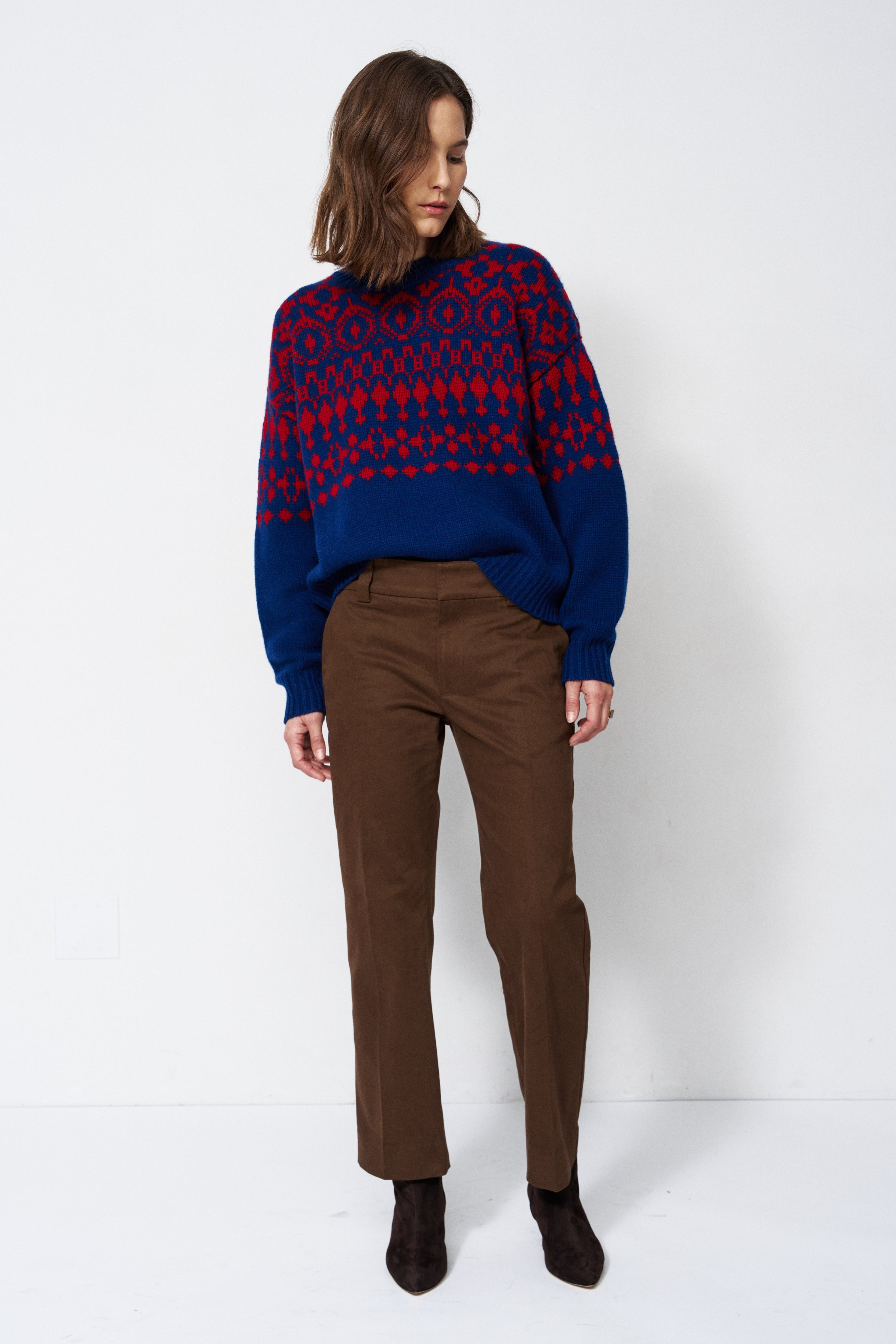 A woman stands against a plain white studio background wearing a navy and red patterned knit sweater, brown trousers, and dark ankle boots.