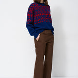 A woman stands against a plain white studio background wearing a navy and red patterned knit sweater, brown wide-leg trousers, and dark pointed ankle boots.