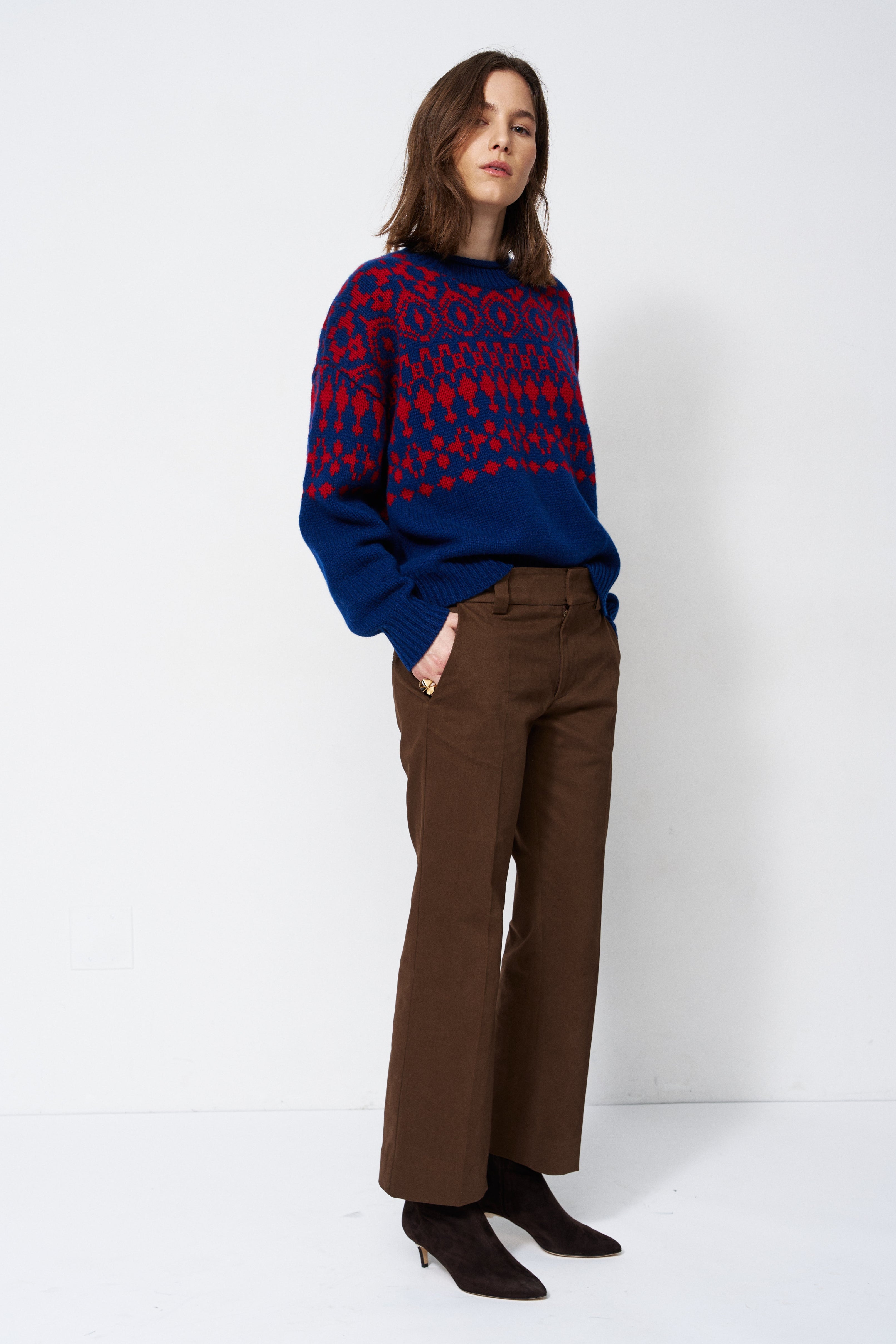 A woman stands against a plain white studio background wearing a navy and red patterned knit sweater, brown wide-leg trousers, and dark pointed ankle boots.