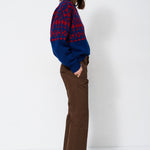 A woman stands in profile wearing a blue and red patterned knit sweater, brown wide-leg trousers, and dark brown pointed-toe heels against a plain white studio background.