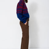 A woman stands in profile wearing a blue and red patterned knit sweater, brown wide-leg trousers, and dark brown pointed-toe heels against a plain white studio background.