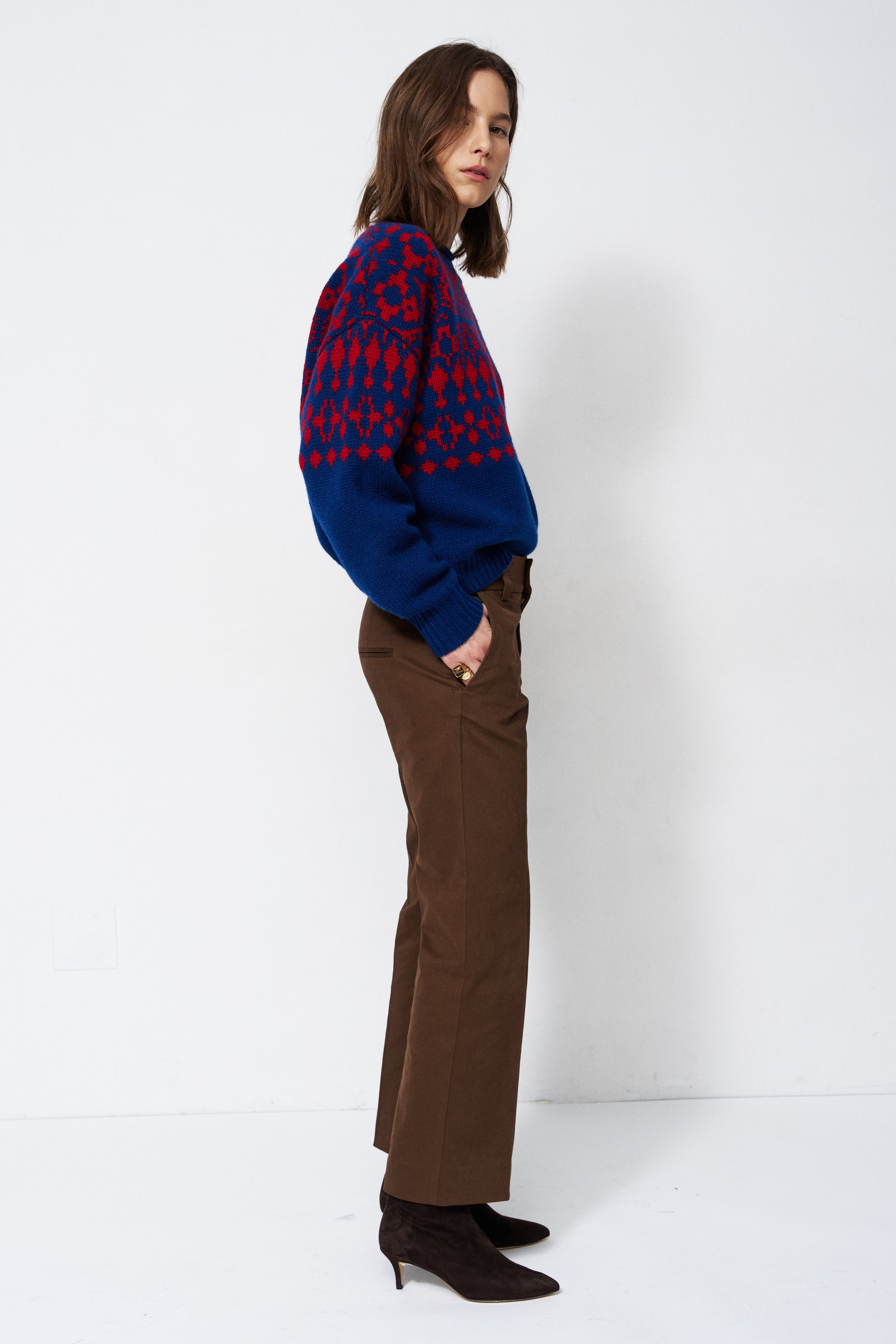A woman stands in profile wearing a blue and red patterned knit sweater, brown wide-leg trousers, and dark brown pointed-toe heels against a plain white studio background.