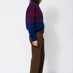 A woman stands in profile wearing a blue and red patterned knit sweater, brown wide-leg trousers, and black pointed-toe heels against a plain white studio background.