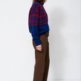 A woman stands in profile wearing a blue and red patterned knit sweater, brown wide-leg trousers, and black pointed-toe heels against a plain white studio background.