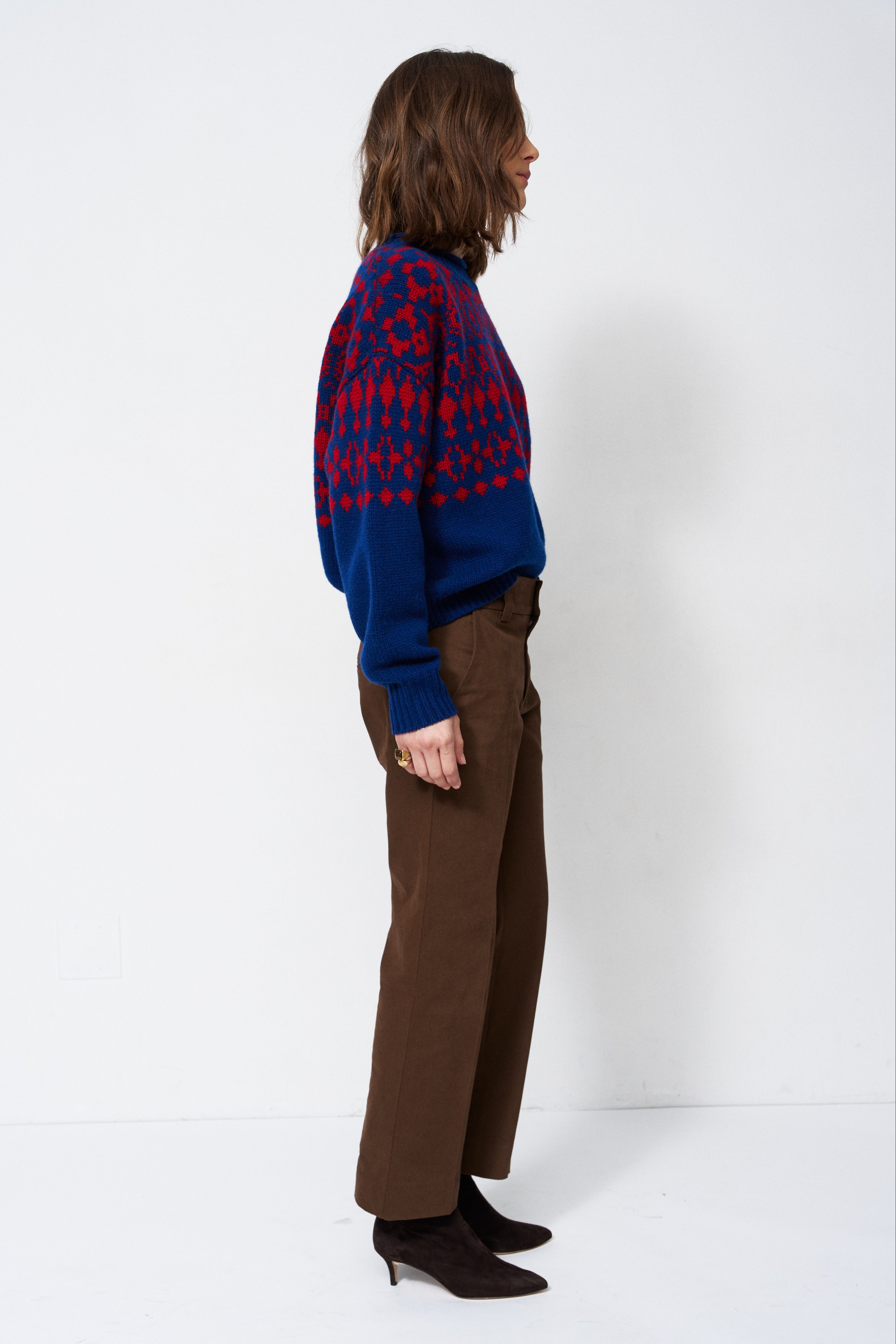 A woman stands in profile wearing a blue and red patterned knit sweater, brown wide-leg trousers, and black pointed-toe heels against a plain white studio background.