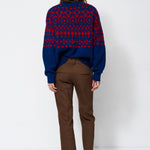 A woman is shown from behind wearing a navy blue and red patterned knit sweater, brown trousers, and black pointed-toe heels against a plain white studio background.
