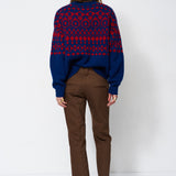 A woman is shown from behind wearing a navy blue and red patterned knit sweater, brown trousers, and black pointed-toe heels against a plain white studio background.