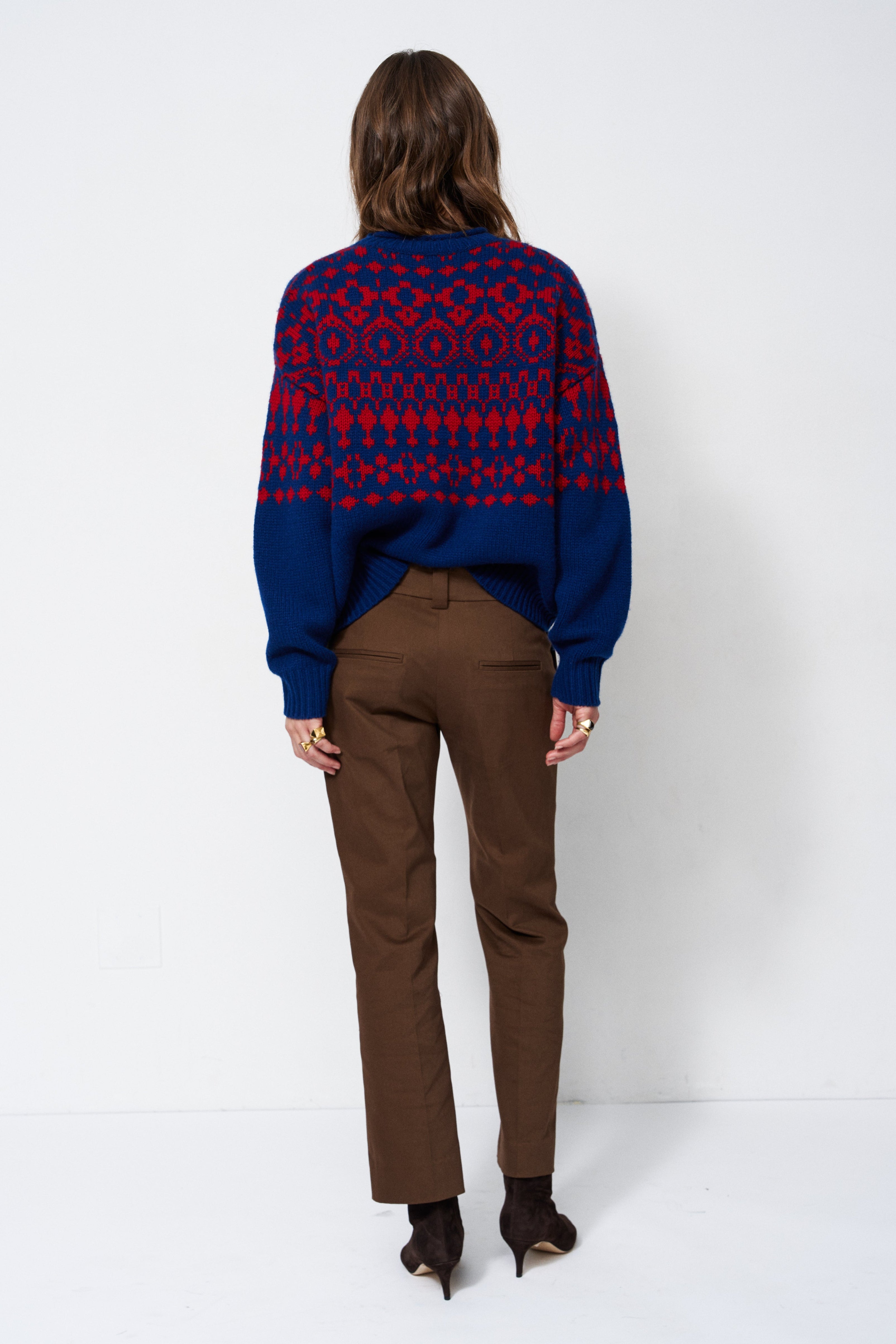 A woman is shown from behind wearing a navy blue and red patterned knit sweater, brown trousers, and black pointed-toe heels against a plain white studio background.