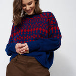 A woman wearing a navy blue and red patterned knit sweater with brown trousers, standing against a light gray studio background.