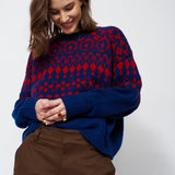 A woman wearing a navy blue and red patterned knit sweater with brown trousers, standing against a light gray studio background.