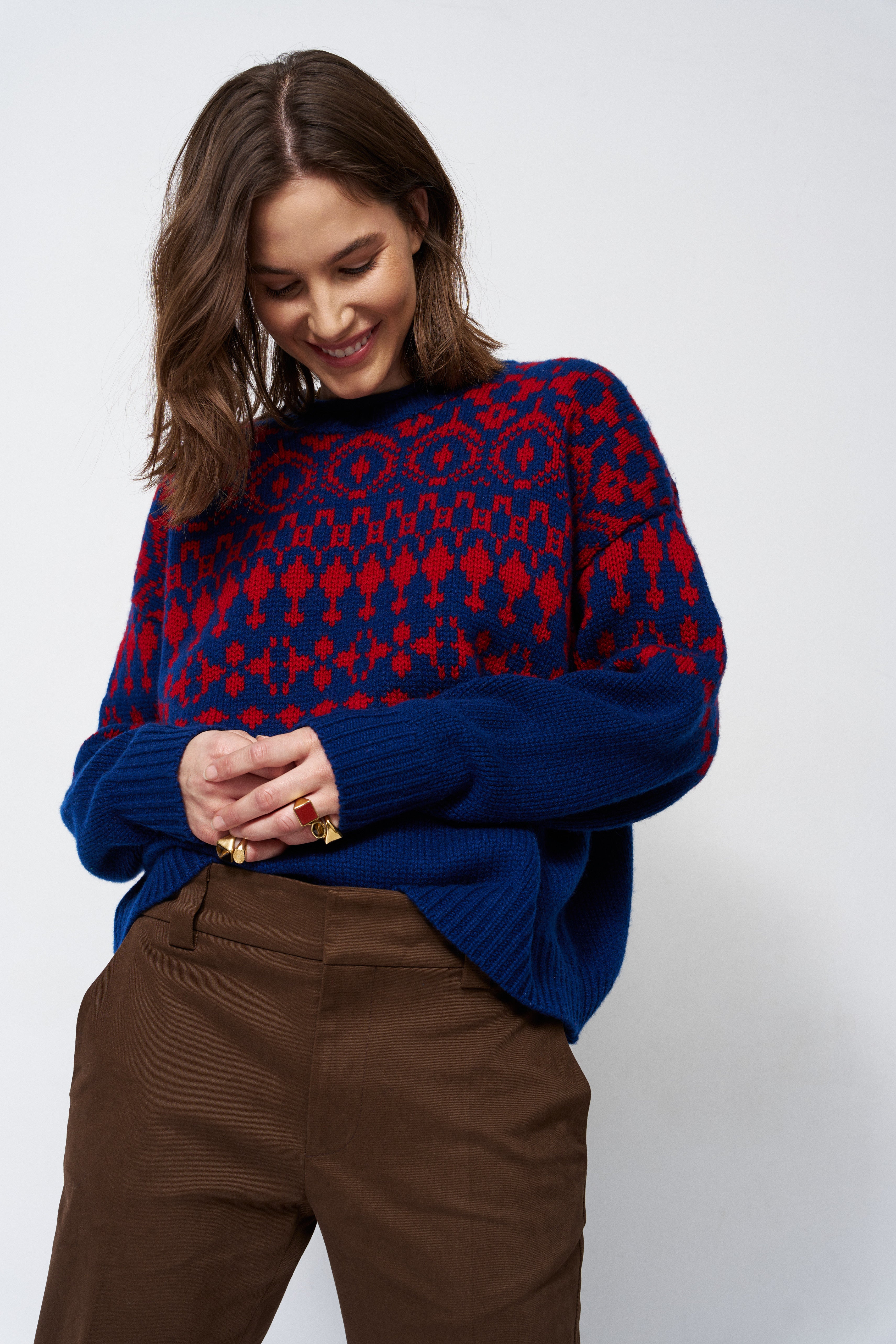 A woman wearing a navy blue and red patterned knit sweater with brown trousers, standing against a light gray studio background.