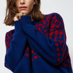 A woman wearing a navy blue knit sweater with a red geometric pattern, styled with brown trousers and gold rings against a light gray background.