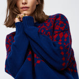 A woman wearing a navy blue knit sweater with a red geometric pattern, styled with brown trousers and gold rings against a light gray background.