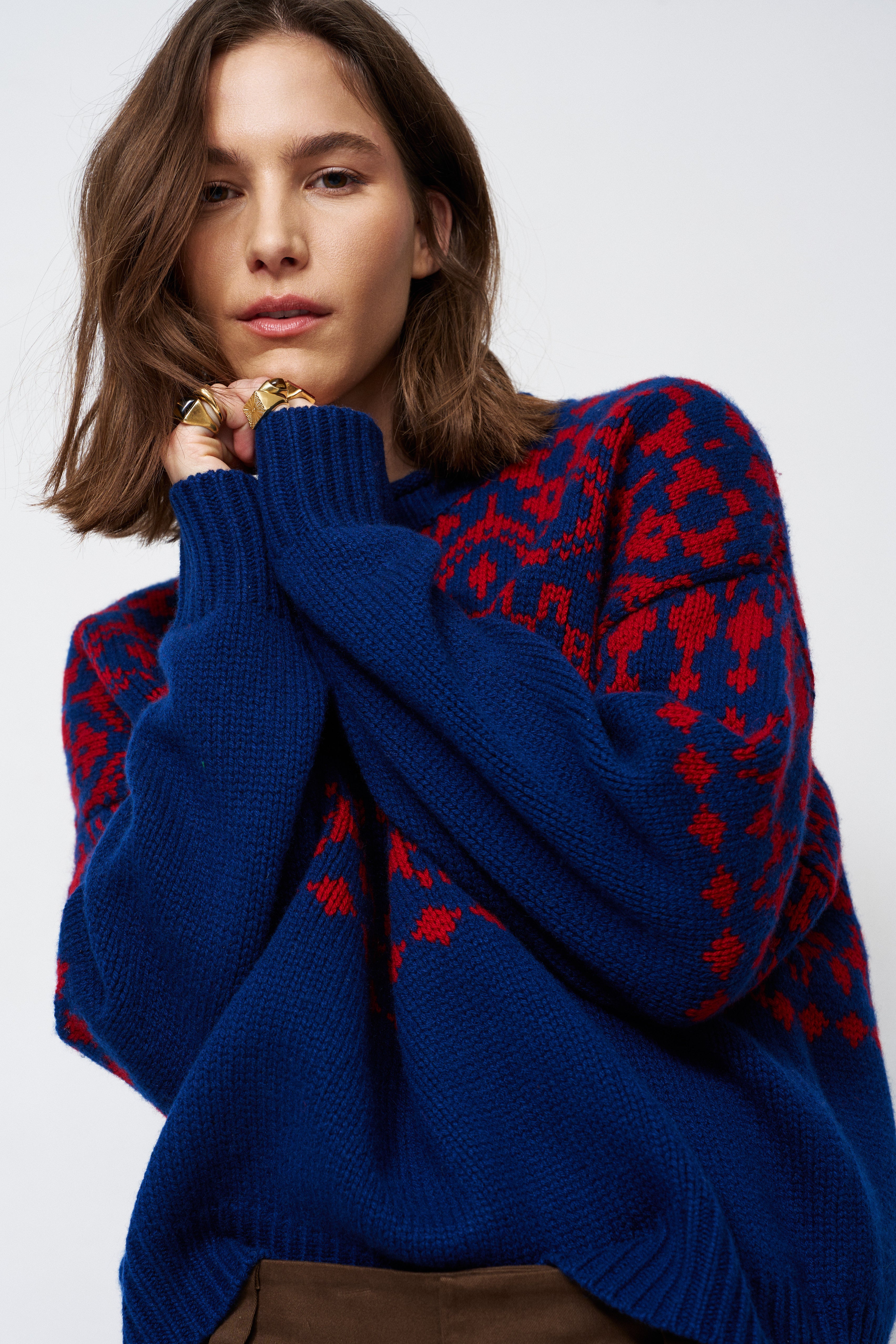 A woman wearing a navy blue knit sweater with a red geometric pattern, styled with brown trousers and gold rings against a light gray background.