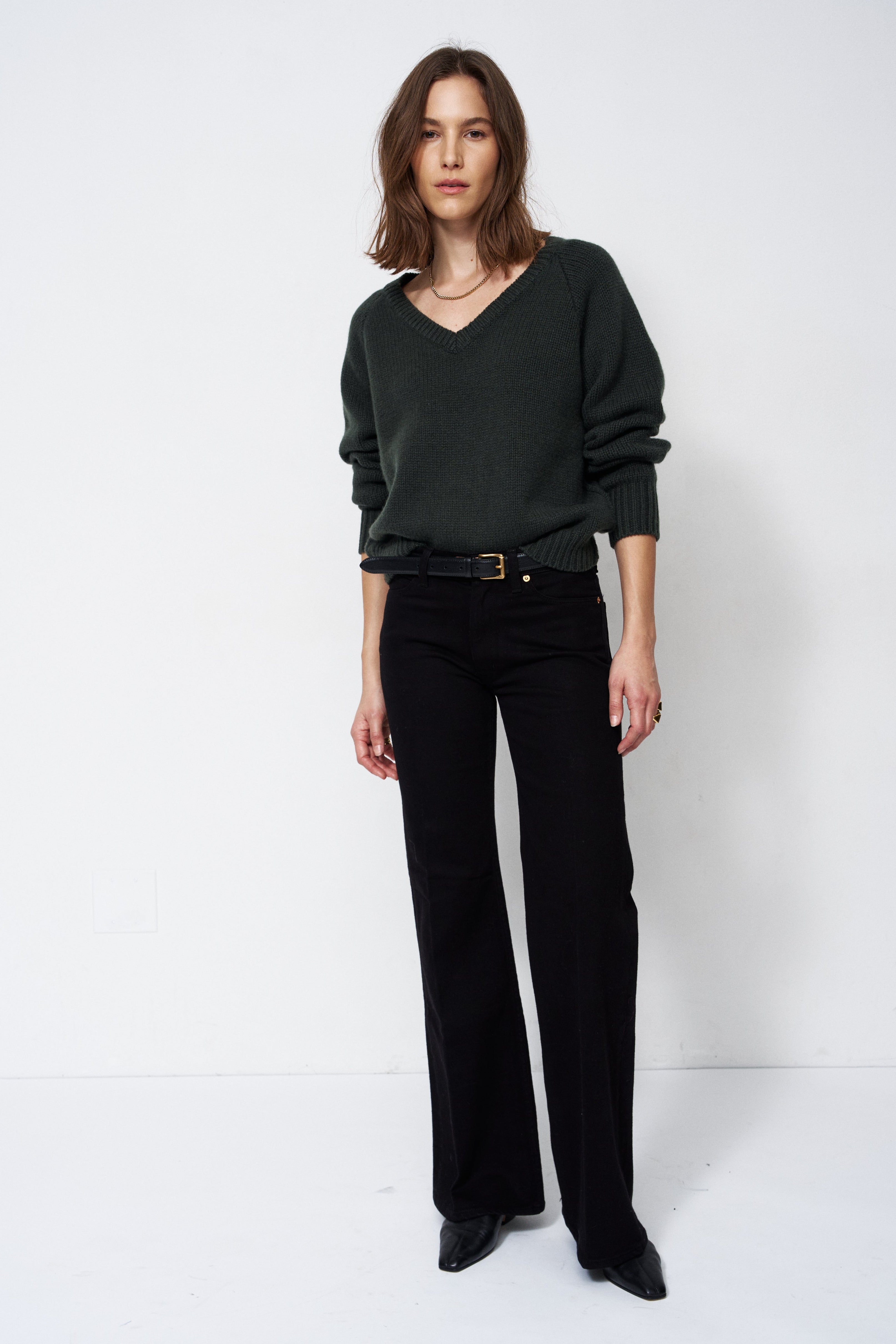 A woman modeling a dark green V-neck knit sweater and black wide-leg trousers with a black belt, standing against a plain white studio background.