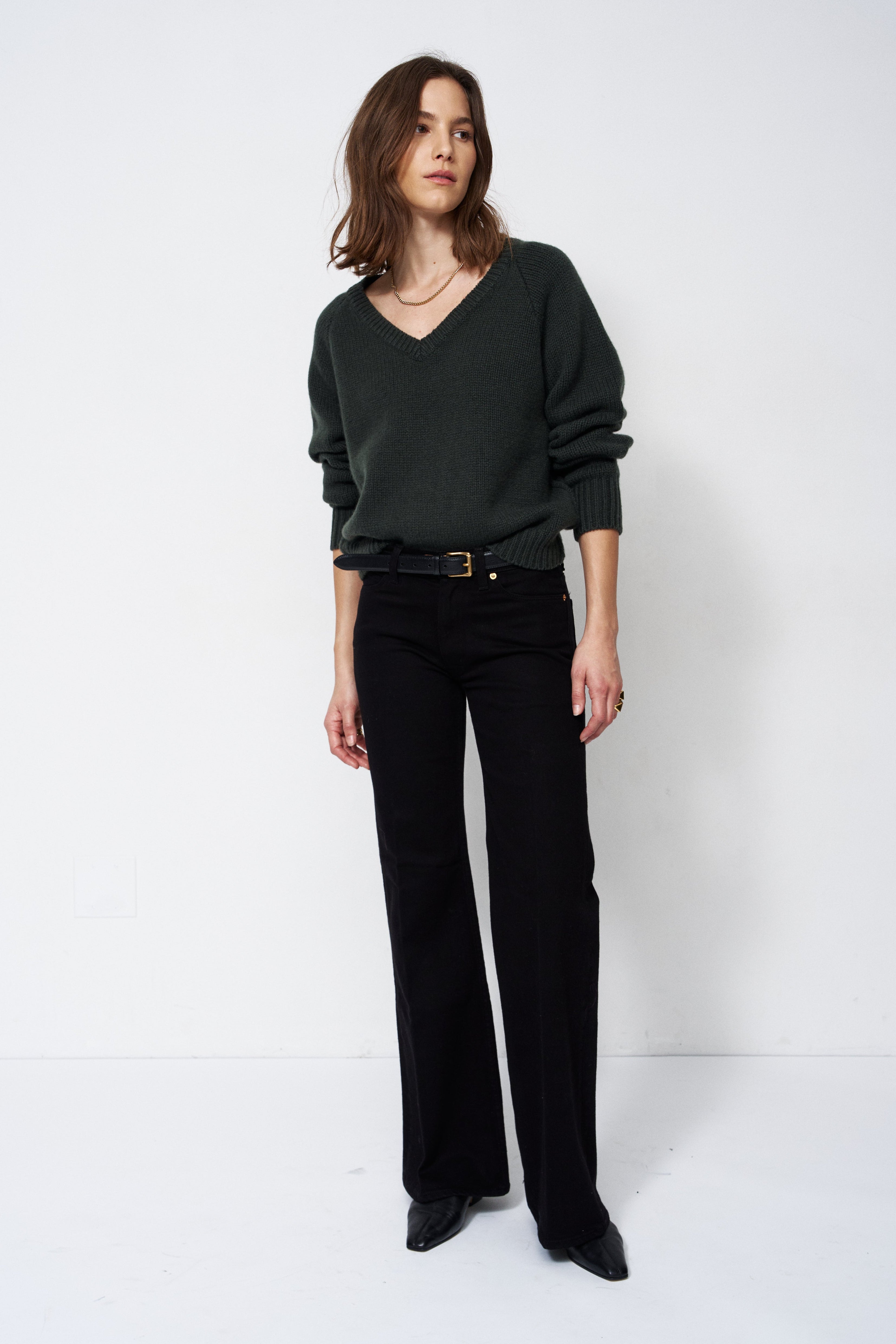 A woman modeling a dark green V-neck knit sweater and black wide-leg trousers with a black belt, standing against a plain white studio background.