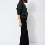 A woman stands in profile wearing a dark green long-sleeve knit sweater and black wide-leg trousers against a plain white studio background.
