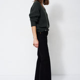 A woman stands in profile wearing a dark green long-sleeve knit sweater and black wide-leg trousers against a plain white studio background.