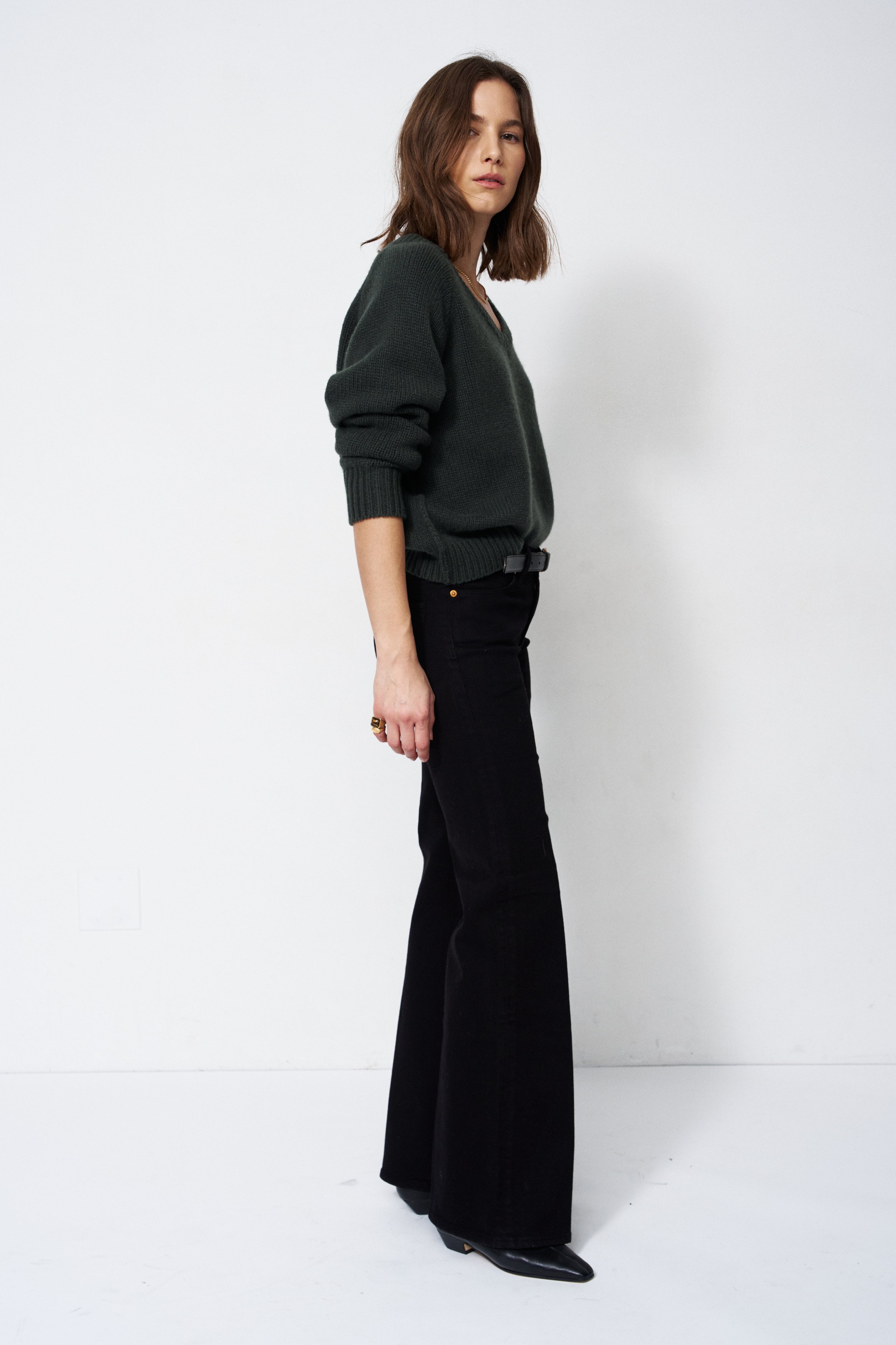 A woman stands in profile wearing a dark green long-sleeve knit sweater and black wide-leg trousers against a plain white studio background.