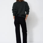 A model viewed from behind wearing a dark green knit sweater and black wide-leg trousers, standing against a plain white studio background.