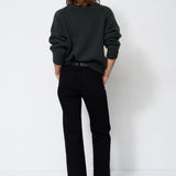 A model viewed from behind wearing a dark green knit sweater and black wide-leg trousers, standing against a plain white studio background.