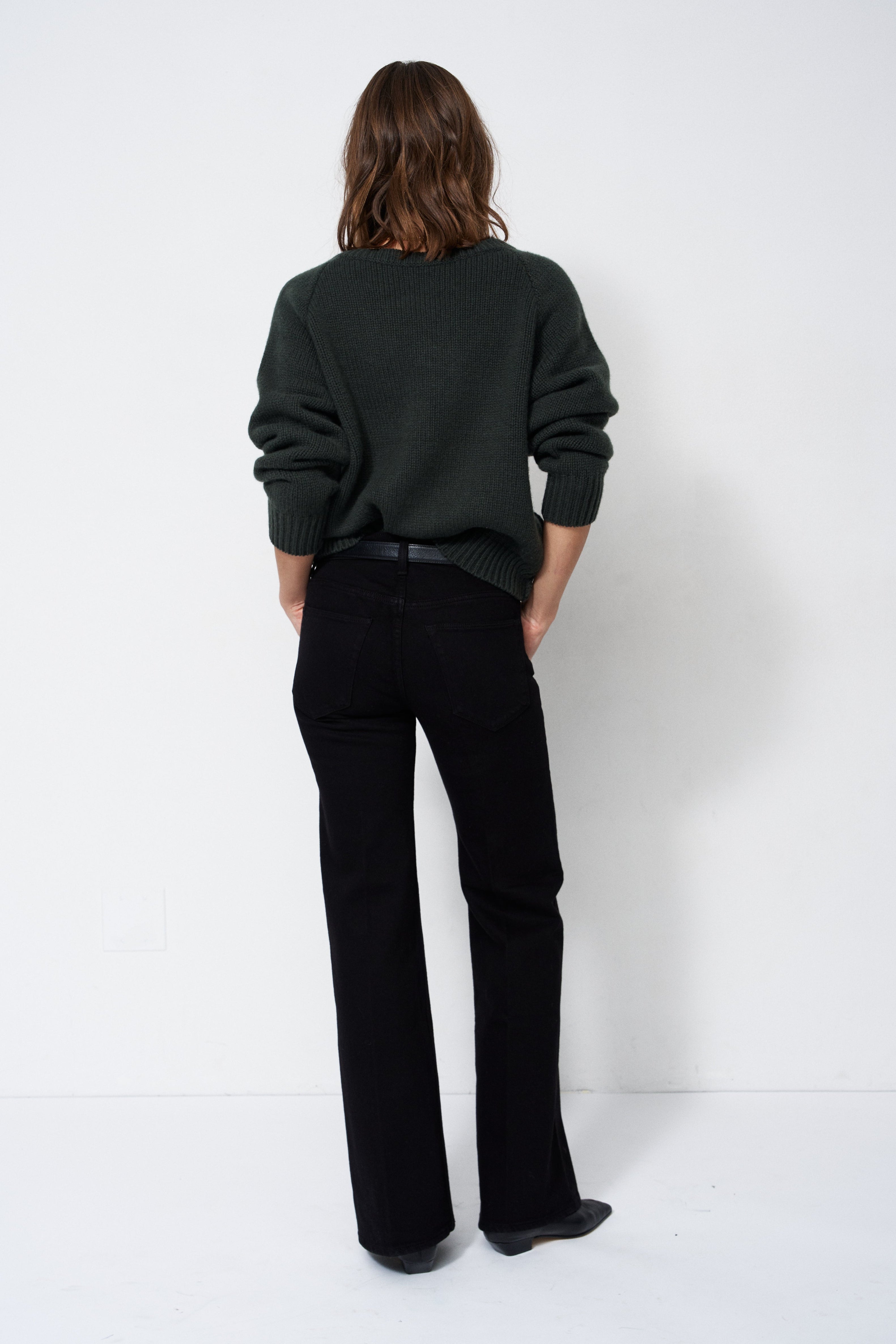 A model viewed from behind wearing a dark green knit sweater and black wide-leg trousers, standing against a plain white studio background.
