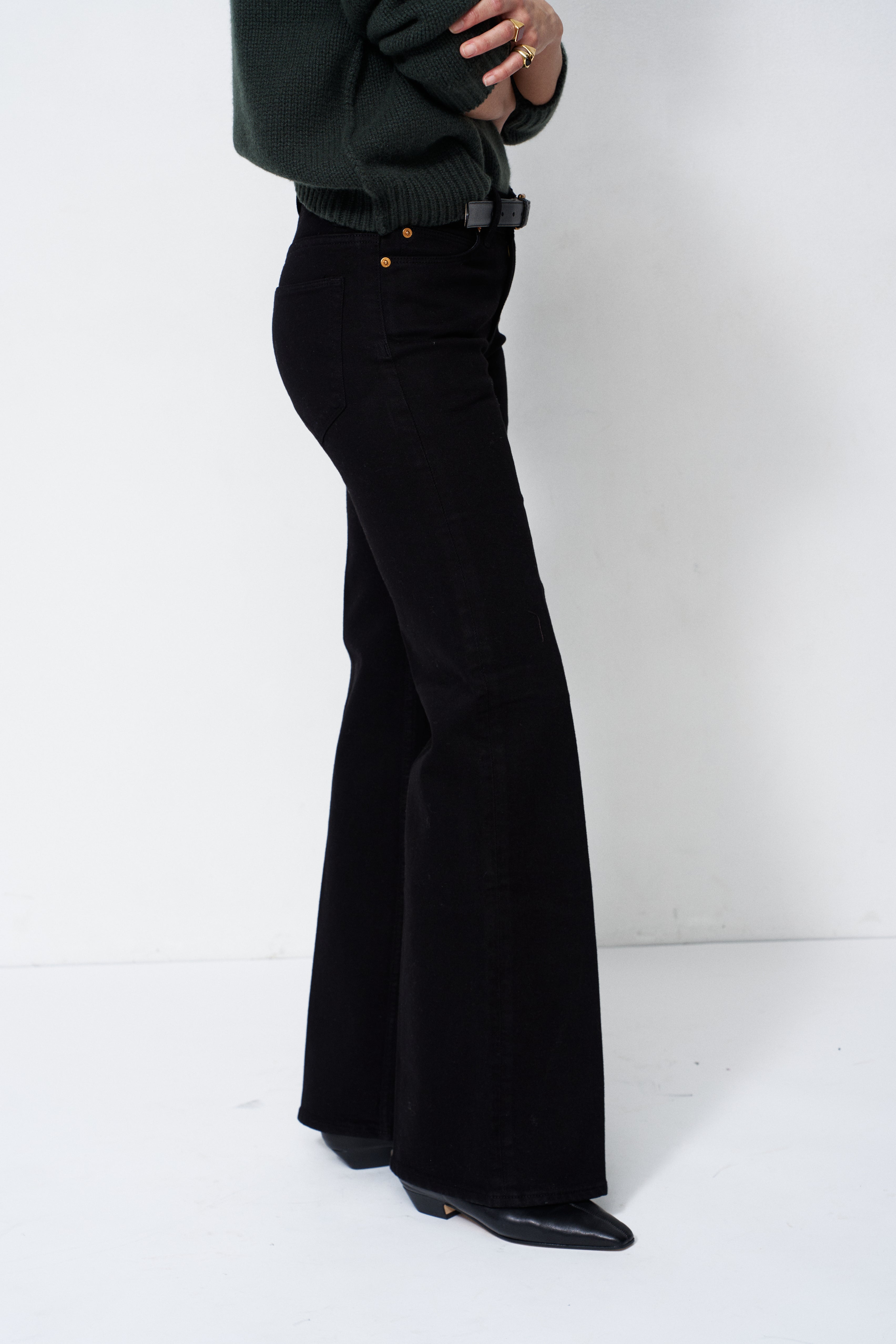 A person wearing black wide-leg flared pants, a dark green sweater, and black pointed shoes, shown from the waist down against a plain white studio background.