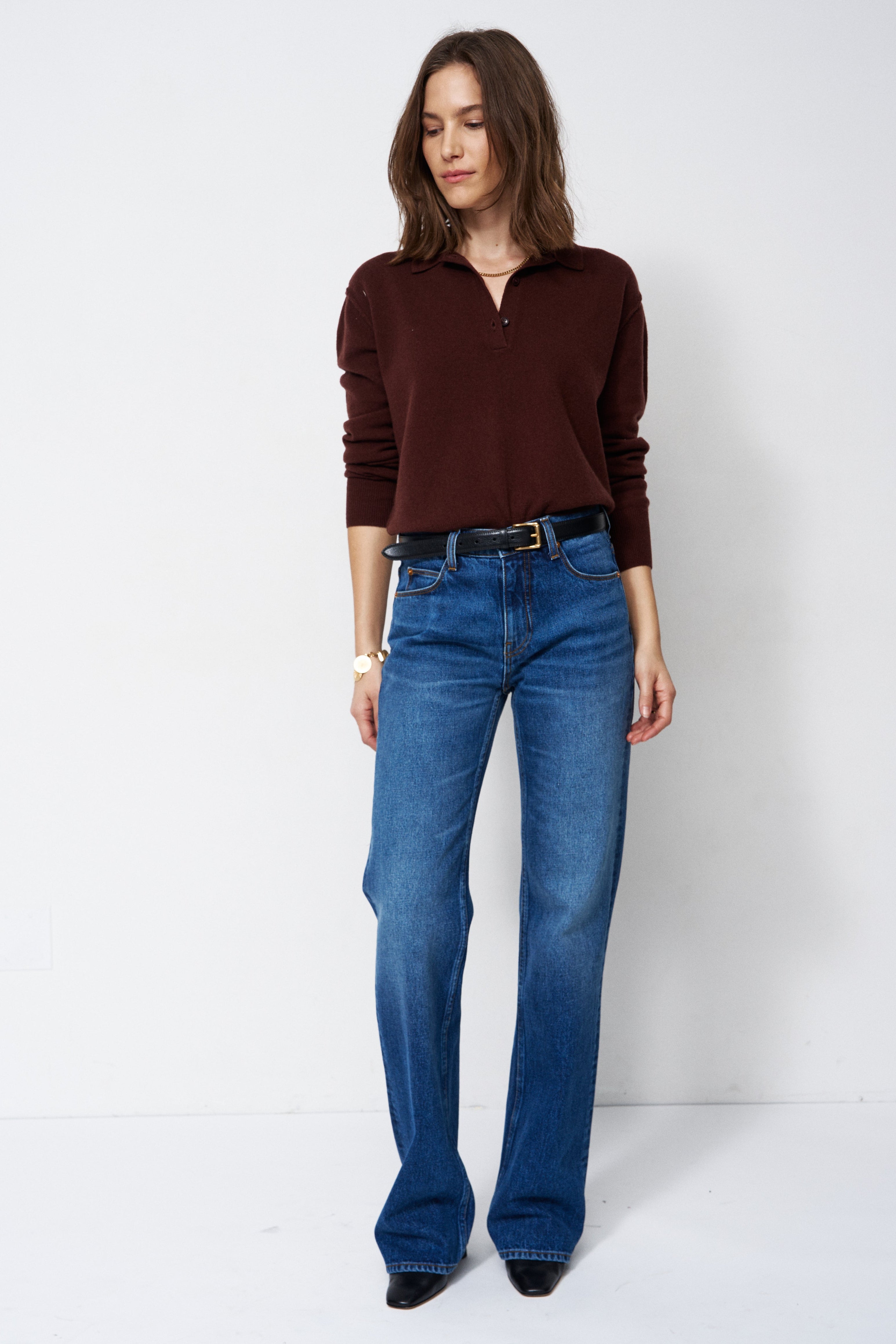 A woman modeling a dark brown long-sleeve polo-neck knit top and blue straight-leg jeans against a plain white studio background.