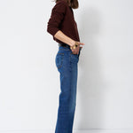 A woman modeling a pair of high-waisted, wide-leg blue jeans paired with a dark brown long-sleeve top, standing in profile against a plain white studio background.