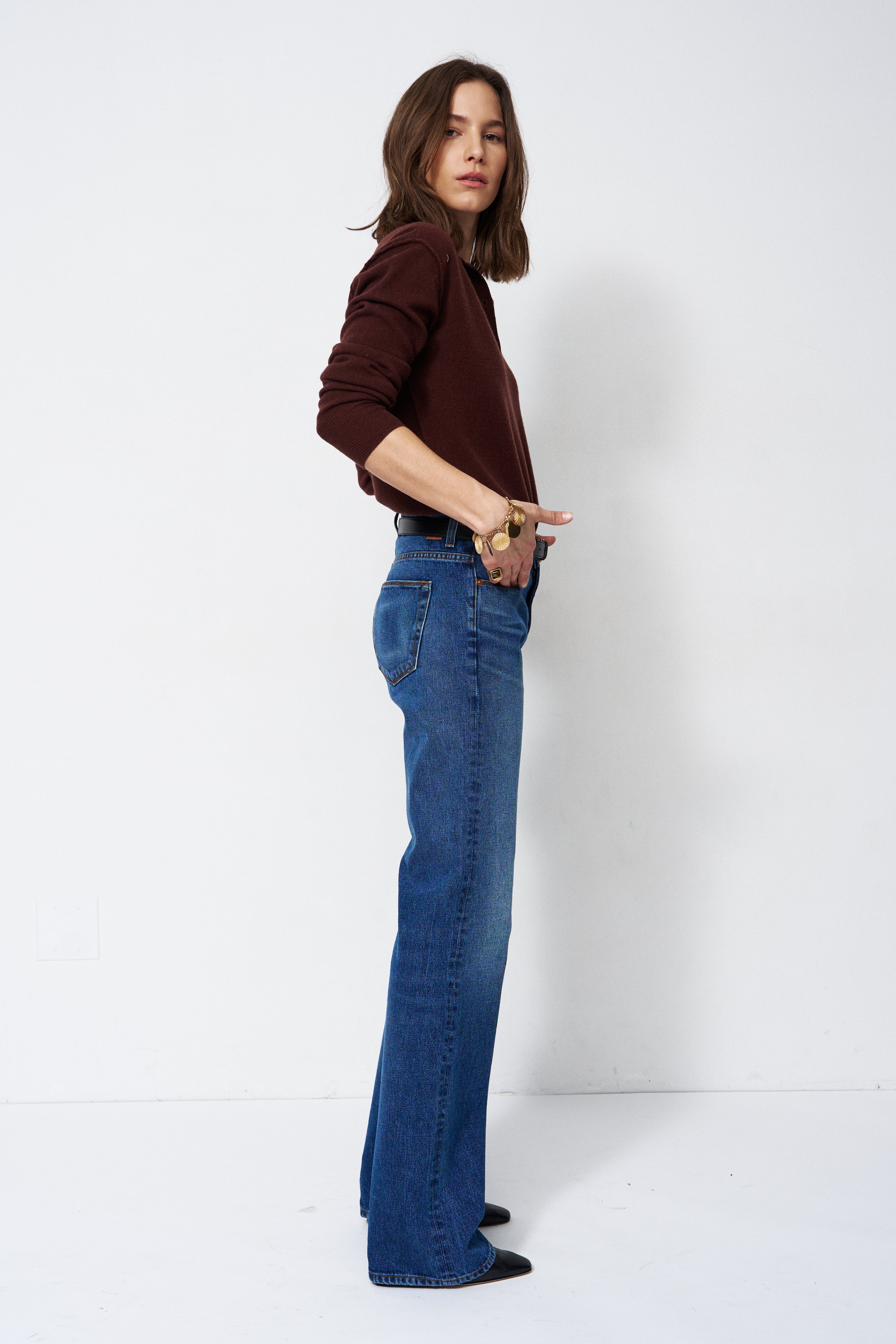 A woman modeling a pair of high-waisted, wide-leg blue jeans paired with a dark brown long-sleeve top, standing in profile against a plain white studio background.