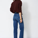 A woman is shown from behind wearing blue flare jeans and a dark brown long-sleeve top, standing against a plain white studio background.