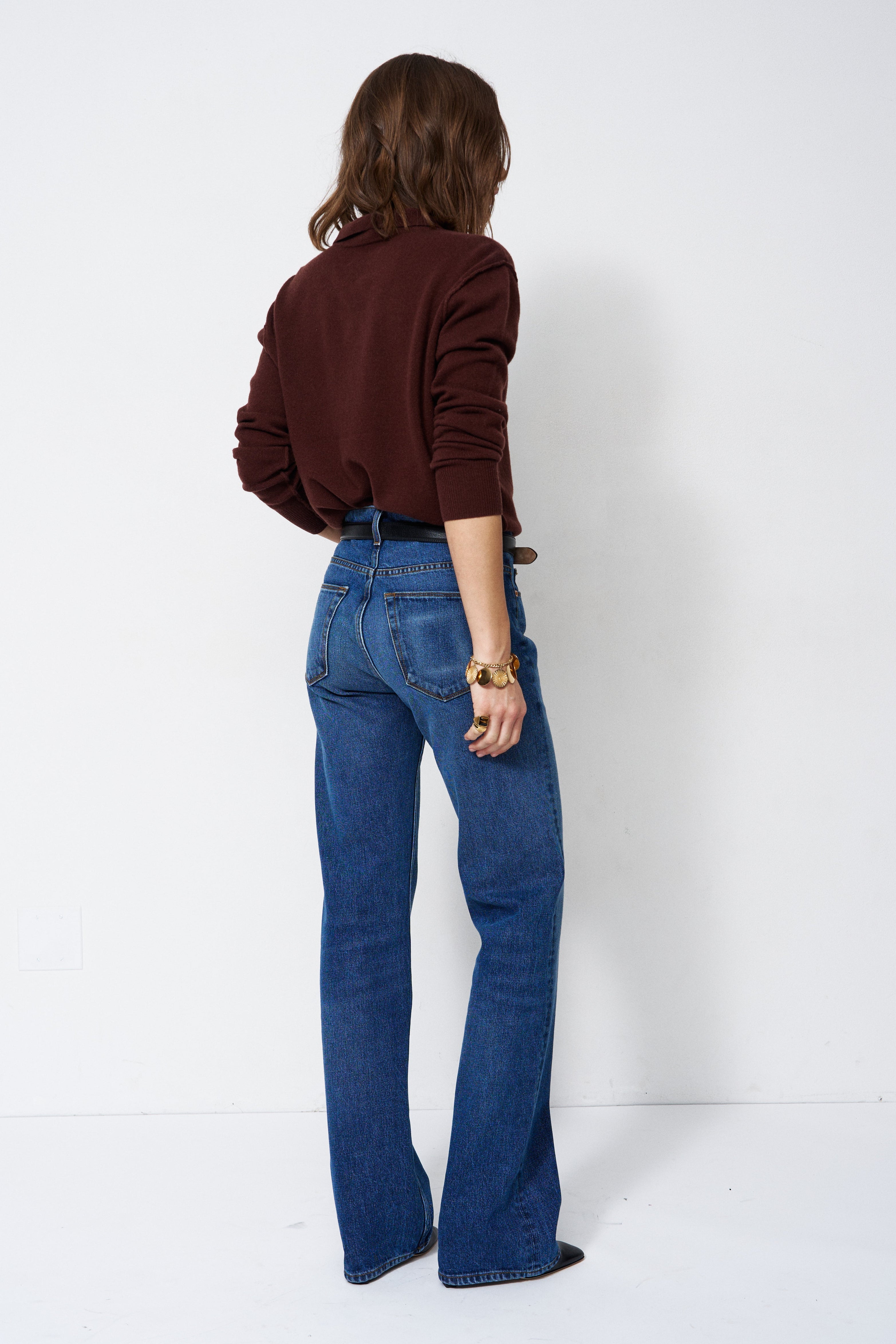 A woman is shown from behind wearing blue flare jeans and a dark brown long-sleeve top, standing against a plain white studio background.