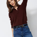 A woman modeling a dark brown long-sleeve polo shirt with blue jeans and a black belt, posing against a light gray studio background.