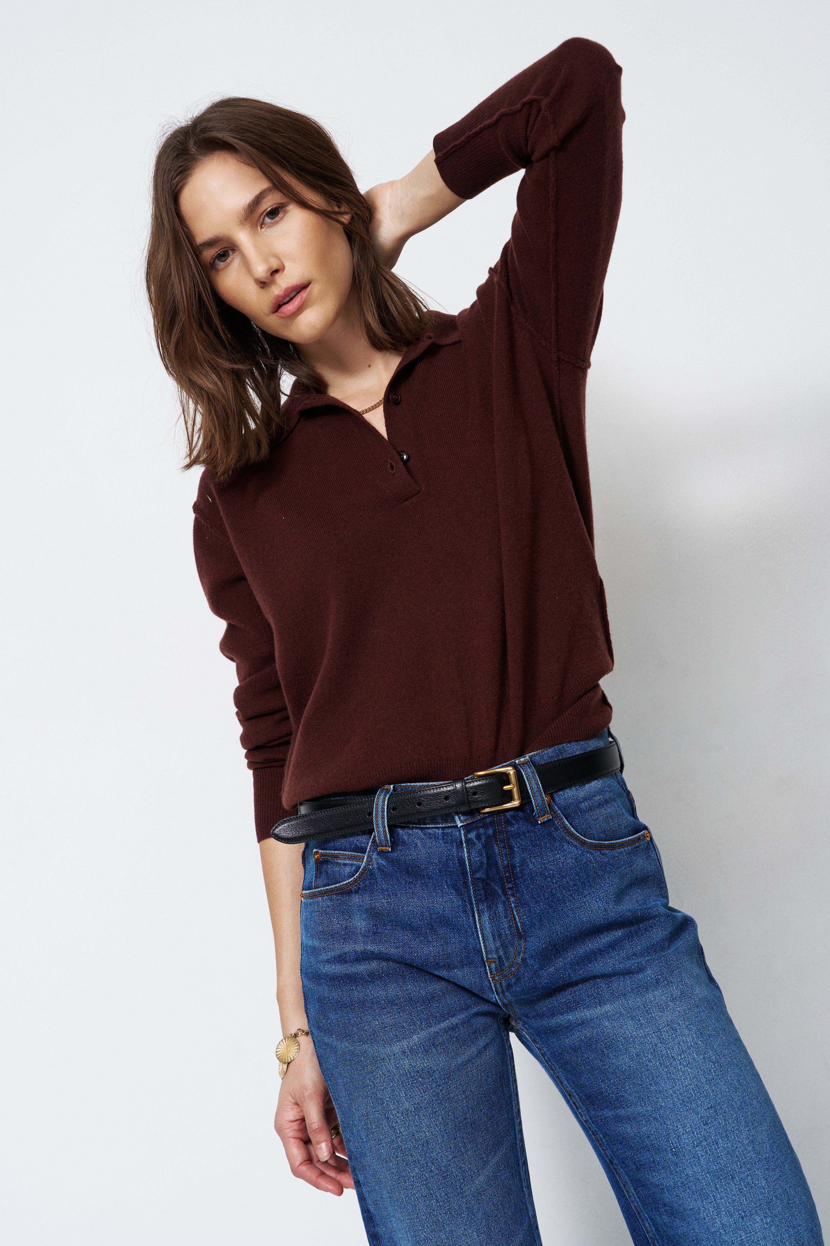 A woman modeling a dark brown long-sleeve polo shirt with blue jeans and a black belt, posing against a light gray studio background.