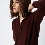 A woman wearing a dark brown long-sleeve knit polo shirt with a partial button placket, paired with blue jeans, photographed against a clean white studio background.